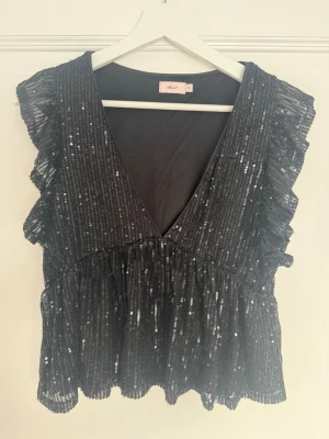 Svart glittrig topp - Säljer en supersnygg svart festtopp från Nelly med glittriga paljetter och volangdetaljer. Toppen har v-ringning och är ärmlös💞
