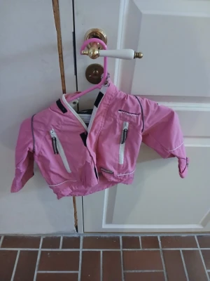 Rosa vindjacka från protech - Snygg rosa vindjacka från protech med vita dragkedjor och reflexdetaljer. Jackan har meshfoder, hög krage och två fickor framtill. Perfekt för blåsiga dagar och enkel att matcha med andra plagg.