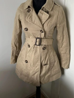 Beige trenchcoat med bälte - Stilren beige trenchcoat med dubbelknäppning och bälte i midjan. Figursydd passform och snygga detaljer. Perfekt nu till våren! Xs/S. Inga defekter bara testad