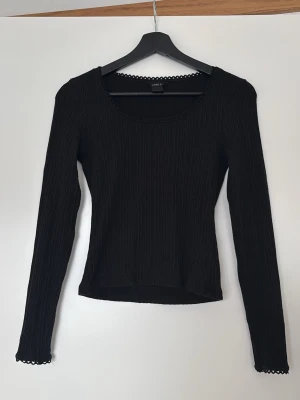 Svart ribbad topp från Lindex - Säljer en svart långärmad topp från Lindex med ribbad struktur och rundad halsringning dekorerad med spetskant. Toppen har en slim passform och är perfekt för att styla med jeans eller kjol.