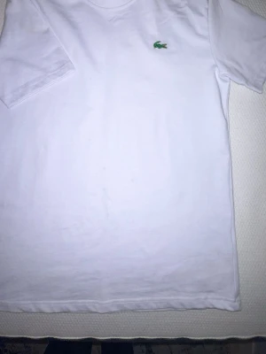 Vit t-shirt från Lacoste - Klassisk vit t-shirt från Lacoste med den ikoniska gröna krokodilloggan broderad på bröstet. T-shirten har rund halsringning och korta ärmar. Tillverkad i mjuk bomull för en skön och avslappnad stil.