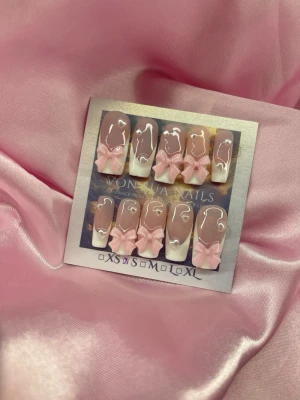 PinkBows (S);Medium Square - Snygga handgjorda press-on naglar i en mjuk nude färg med vit fransk tipp och 3D rosa rosetter. Naglarna har en glansig finish och en rak, något fyrkantig form. Perfekt för dig som vill ha en söt och trendig look.