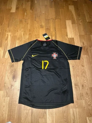 Portugal away kit 2006 X C.Ronaldo 17 - Snygg svart Portugal matchtröja från Nike med gula detaljer och nummer 17. På bröstet finns Portugals landslagsemblem och på ryggen står det C. Ronaldo med stort gult nummer. Tillverkad i lätt och ventilerande material, perfekt för fotboll eller gymmet.