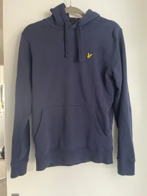 Mörkblå hoodie från Lyle & Scott - Snygg mörkblå hoodie från Lyle & Scott med klassisk gul logga på bröstet. Hoodien har huva med dragsko, stor magficka och ribbade muddar vid ärmslut och nederkant. Tillverkad i mjuk bomullsblandning som är skön att bära.