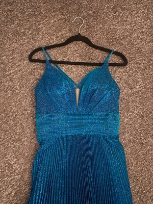 Balklänning - Säljer en blå glittrig balklänning med smala axelband och djup v-ringning i storlek 34. Använd 1 gång. 