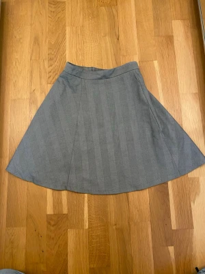 Grå kjol med diskreta ränder - Snygg grå kjol med subtilt randigt mönster. Kjolen har en lätt utställd form och är tillverkad i ett mjukt tyg som känns bekvämt. Perfekt att matcha med en enkel topp eller stickad tröja för en stilren look.