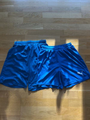 Blåa sportshorts från Puma - Två par blåa sportshorts från Puma med elastisk midja och klassisk logga på ena benet. Perfekta för träning eller fotboll, tillverkade i lätt och snabbtorkande syntetmaterial. Snygg enkel design utan mönster.