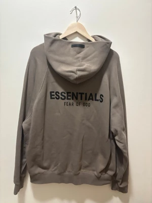 Brun hoodie Essentials Fear of God - Säljer en brun hoodie från Essentials Fear of God med stor svart logga på ryggen. Hoodien har en klassisk huva och är gjord i mjukt, fleecefodrat material som känns riktigt skönt. Perfekt för dig som gillar streetwear och vill ha en clean look.