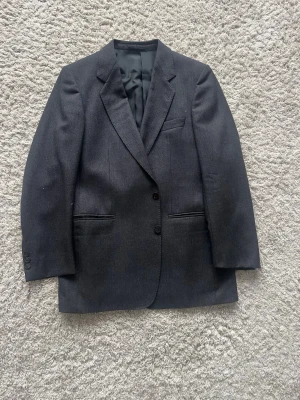 Mörkgrå kavaj från Theowormland - Stilren mörkgrå kavaj från Theoeormland med två knappar framtill, klassisk krage och tre fickor. Kavajen har en rak passform och är helfodrad med svart innerfoder. Perfekt för en dressad look.