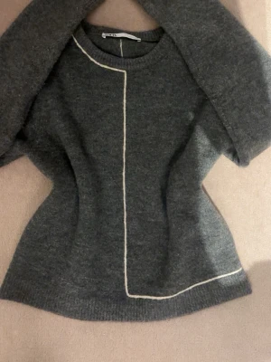 Mörkgrå stickad tröja med vit detalj - Säljer en mörkgrå stickad tröja med en unik vit linje som går längs framsidan och runt nederkanten. Tröjan är gjord i mjukt material som känns skönt mot huden. 