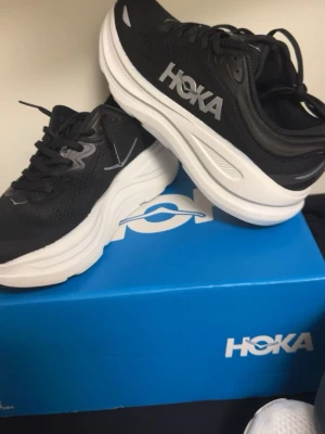 Svarta Hoka sneakers med vit sula - Snygga svarta sneakers från Hoka med vit, tjock sula och ventilerande mesh-ovandel. Skorna har klassisk snörning och Hoka-logga i vitt på sidan. Perfekta för dig som gillar sportig och modern stil.