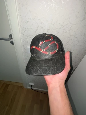Gucci keps med orm och GG-mönster - Säljer en svart Gucci keps med klassiskt GG-mönster och en stor röd och vit orm broderad framtill. Kepsen har böjd skärm och mesh på sidorna för extra ventilation. Perfekt för dig som vill sticka ut med en lyxig och trendig accessoar.