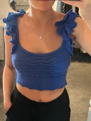 Blå stickad croppad linnetopp - Supersöt blå croppad linnetopp med volangdetaljer på axelbanden. Toppen är stickad med vågigt mönster och har en tajt passform som framhäver midjan. Perfekt för dig som vill ha en trendig och unik topp till sommaren.