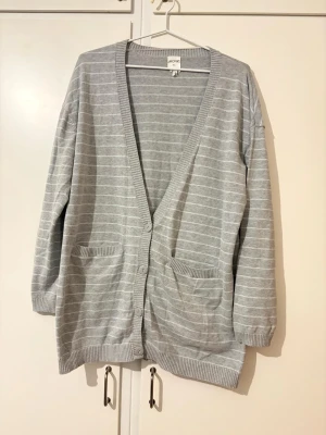 Grå randig kofta från Monki - Säljer en grå kofta från Monki med tunna vita ränder. Koftan har djup V-ringning, knäppning med fyra knappar och två fickor framtill. Materialet är mjukt . Ribbstickade muddar vid ärmslut och nederkant.