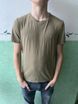 Beige t-shirt med liten logga - En enkel beige t-shirt i bomull med rund hals och korta ärmar. Liten diskret logga på bröstet. Perfekt basplagg för en avslappnad stil.