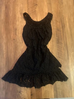 Svart spetsklänning med volang - Snygg svart klänning i spets med volang. Klänningen har breda axelband och ett feminint snitt som framhäver midjan. Perfekt för dig som gillar romantiska detaljer och en klassisk look.