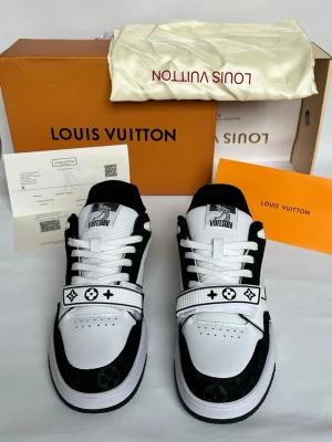 Louis Vuitton svartvita sneakers - Snygga svartvita sneakers från Louis Vuitton med klassiska monogramdetaljer på remmen och hälen. Skorna har en låg silhuett, vita snören och meshpaneler på sidorna för extra stil. Perfekta för dig som vill sticka ut med exklusiva detaljer och modern design.