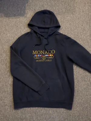 Marinblå Monaco Monte-Carlo hoodie - Snygg marinblå hoodie med huva i storlek M. På bröstet står det 'MONACO MONTE-CARLO' i guld text med broderade flaggor och ett ankare. Går endast att köpa lokalt i Monaco. Nypris 1700, mitt pris 399. Bara att höra av sig vid frågor! Pris går alltid att diskutera 🙌