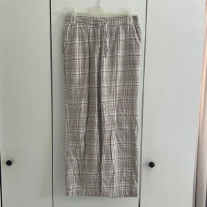 Beiga pyjamasbyxor - Säljer ett par beige pyjamasbyxor med rutigt mönster i vitt och grått. Byxorna har rak passform och resår i midjan för extra komfort. Perfekta för mysiga hemmakvällar eller lata morgnar.