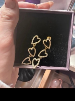 Guldiga örhängen med hjärtan - Säljer ett par guldplätterade statement örhängen i guldton med flera sammankopplade hjärtan. Varje hjärta är dekorerat med små gnistrande stenar som ger extra bling. Perfekta för dig som vill sticka ut och addera lite glamour till din look.