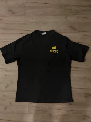 Extremt sällsynt Svart NOCCO t-shirt med tryck - Svart NOCCO-t-shirt i mjuk bomull med gul logga på bröstet och stort “TIME TO FOCUS”-tryck på ryggen. Extremt sällsynt modell som producerats i mycket begränsat antal – finns endast ett fåtal exemplar. Ett unikt plagg för samlare och NOCCO-fans. BUDGIVNING AKTIV, PRIS GÅR ATT DISKUTERA