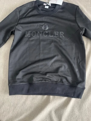 Mörkgrå sweatshirt från Moncler - Snygg mörkgrå sweatshirt från Moncler med broderad logotyp och text på bröstet. Tröjan har rund halsringning, ribbade muddar vid ärmslut och nederkant samt en stilren design. Perfekt för dig som gillar exklusiva märken och enkel stil.
