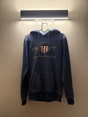 Blå hoodie från GANT med tryck - Mörkblå hoodie från GANT med broderad logga och texten 'American Sportswear' på bröstet. Klassisk huva med dragsko och stor magficka framtill. Tillverkad i mjukt bomullsmaterial, perfekt för en avslappnad och sportig stil.