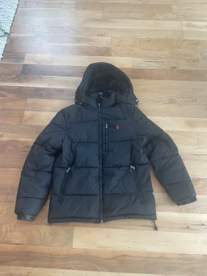 Svart pufferjacka från Polo Ralph Lauren - Säljer en svart pufferjacka från Polo Ralph Lauren med röd broderad logga på bröstet. Jackan har huva, dragkedja framtill, två sidofickor med dragkedja och en bröstficka. Perfekt för kallare dagar och riktigt skön att bära.