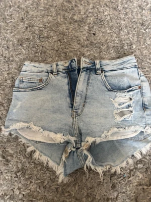 Ljusblå denimshorts med fransar - Säljer ett par ljusblå denimshorts med hög midja och råa fransade kanter. Shortsen har slitningar framtill, klassiska fem fickor och dragkedja med knapp. Perfekta för sommaren och ger en avslappnad vibe.