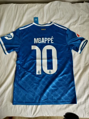 Blå Real Madrid Mbappé matchtröja - Snygg blå Real Madrid matchtröja från Adidas med Mbappé och nummer 10 på ryggen. Tröjan har vita detaljer, klubbmärke och sponsortryck på bröstet samt klassiska Adidas-ränder på axlarna. Tillverkad i lätt och ventilerande material, perfekt för fotboll eller träning.