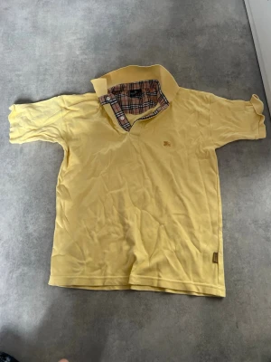 Gul kortärmad piké från Burberry - Säljer en ljusgul kortärmad pikétröja från Burberry med klassiskt rutigt mönster på insidan av kragen och diskret broderad logga på bröstet. Tillverkad i mjuk bomull och har normal passform. Perfekt för dig som gillar stilrena detaljer.