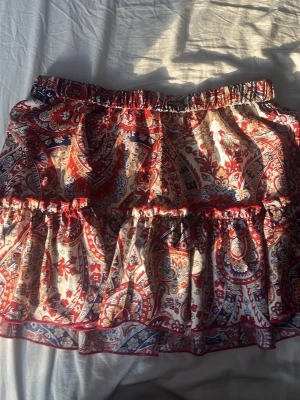 Kort kjol  - Kortkjol med färgstarkt paisleymönster i rött, blått och beige. Kjolen har volangdetaljer och resår i midjan för en bekväm passform. Perfekt för dig som gillar boho-vibbar och vill ha något unikt i garderoben.
