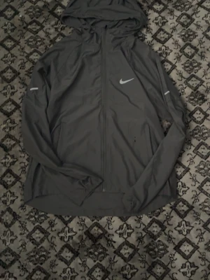 Grå löparjacka med huva från Nike - Snygg grå löparjacka från Nike med huva och dragkedja framtill. Jackan har reflekterande detaljer på ärmarna och Nike-logga på bröstet. Perfekt för löpning eller träning utomhus. Tillverkad i lätt och funktionellt material.