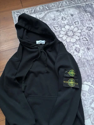 Svart hoodie från Stone Island - Svart hoodie från Stone Island med klassisk huva och dragsko. På vänster ärm sitter två avtagbara Stone Island-logopatchar i svart med gul och grön brodyr. Hoodien har känguruficka framtill och är gjord i mjukt bomullsmaterial. Perfekt för en grisch look. 2 st badkar kommer med den sitter litet i storlek och på mig som är 174 så är den för liten kom gärna med bud.