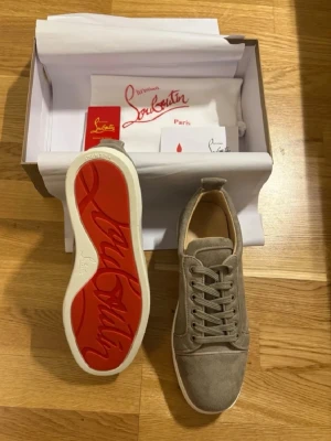 Beige sneakers från Christian Louboutin - Stilrena beige sneakers i mocka från Christian Louboutin med den ikoniska röda sulan och vita kanter. Klassisk låg modell med snörning och diskreta sömmar. Perfekta för dig som vill ha en lyxig touch på din outfit.