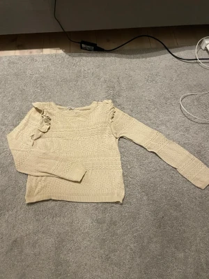 Beige stickad tröja - Snygg beige stickad tröja med volangdetalj på axeln och fina hålmönster över hela plagget. Tröjan har rund halsringning och långa ärmar. Perfekt för dig som gillar en söt och trendig stil.