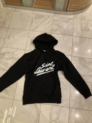 Svart Saint Laurent hoodie - Snygg svart hoodie från Saint Laurent med stor vit logga framtill i handskriftsstil. Hoodien har huva med dragsko och en klassisk magficka. Perfekt för dig som vill ha en stilren och trendig look.