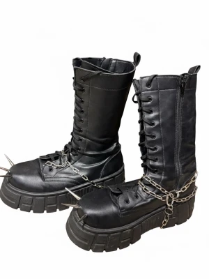 Svarta platåstövlar med kedjor och spikes - Riktigt tunga svarta platåstövlar med snörning, dragkedja på sidan och grov sula. Dekorerade med metallkedjor och stora spikes framtill för en edgy look. Tillverkade i konstläder och perfekta för dig som gillar alternativ stil.