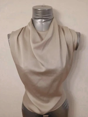 Beige blus  - Stilren beige blus med draperad halsringning och knäppning bak i nacken. Blusen är ärmlös och har en mjuk, något glansig yta som ger ett elegant intryck. Perfekt att matcha med både jeans och kjol för en trendig look.