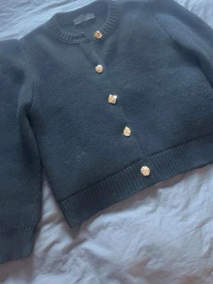 Svart kofta med guldknappar - Stilren svart kofta med långa ärmar. Koftan har fem dekorativa guldknappar framtill. Materialet är stickat och känns mjukt och bekvämt. Perfekt för att lyfta din outfit med en elegant touch. Storlek M men passar mer som en S.