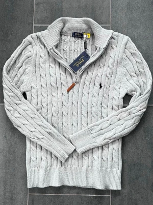 Ljusgrå Polo Ralph Lauren - Snygg ljusgrå kabelstickad tröja från Polo Ralph Lauren med halv dragkedja och ribbad krage. Tröjan har det klassiska broderade logotypen på bröstet och är gjord i mjukt material. Perfekt för dig som gillar stilren och tidlös design.