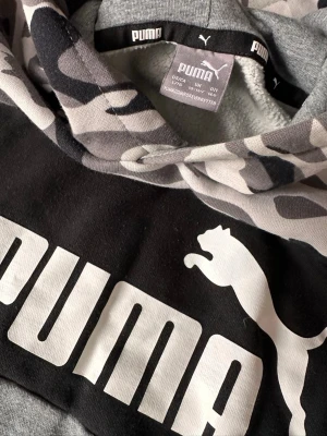 Puma Hoodie 164 Grå camo - Snygg hoodie från Puma i stl 164.  Gtått camo-mönster på huva och ärmar, svart bröstparti med vit Puma-logga och klassisk känguruficka. I fint använt skick.
