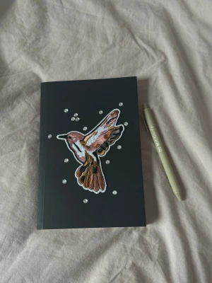 Svart anteckningsbok med kolibri - Snygg svart anteckningsbok med ett broderat motiv av en kolibri i rosa, brunt och vitt på framsidan. Omgiven av små glittriga dekorationer. Perfekt för att skriva ner tankar, idéer eller skisser. Mjukpärm och stilren design.