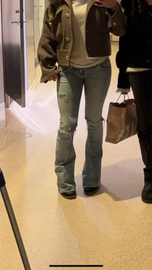 Ljusblå slitna bootcut jeans - Intressekoll! Ett par ljusblå bootcut jeans med slitningar och hål vid knäna samt en mindre reva på låret. Jeansen har klassisk femficksmodell och råa kanter vid knäna för en edgy look. Perfekta för dig som gillar en avslappnad och trendig stil.