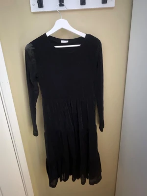 Svart långärmad glitterklänning - En svart långärmad glitter klänning med rund halsringning och lätt rynkad kjol. Klänningen har en enkel och klassisk design som passar till många olika stilar. Materialet ser ut att vara tunt och mjukt, 95% polyamid och 5% Elastan . Klänning är väldigt stretchig så skulle passa en S och kanske M