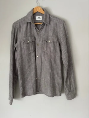 Grå Overshirt från Ciszere - Snygg grå skjorta från Ciszere med tunna vita ränder och klassisk krage. Skjortan har två bröstfickor med knappar och långa ärmar. Tillverkad i ett mjukt material som känns skönt mot huden. Perfekt för en clean och stilren look.