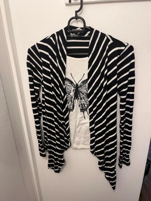 Svartvit randig kofta med draperad front kidsize - Svart och vit långärmad kofta med horisontella och diagonala ränder. Koftan har en öppen, draperad front och längre spetsiga kanter framtill. Tunn och mjuk i materialet, perfekt för lager-på-lager-stil. Kidsize! 146/152