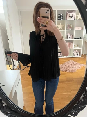 Svart plisserad blus med knyt - Säljer en svart plisserad blus med knyt i halsen. Blusen är ärmlös och har en lös passform som ger ett snyggt fall. Perfekt att styla med jeans eller kjol för en trendig look.