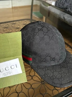 Svart Gucci keps med GG-mönster - Säljer en svart keps från Gucci med klassiskt GG-mönster över hela kepsen. På sidan finns en grön och röd rand som är typisk för Gucci. Materialet är canvas och kepsen har en böjd skärm. Perfekt för dig som vill ha en exklusiv accessoar.
