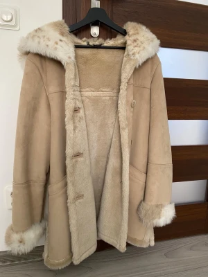 Beige fuskpälsjacka med huva - Snygg beige pälsjacka med mjukt foder och fluffiga detaljer vid huva, ärmslut och nederkant. Jackan har en avslappnad passform, knäppning framtill och stora fickor. Materialet är troligtvis fuskpäls och mockaimitation. Perfekt för dig som vill ha en varm och trendig look.